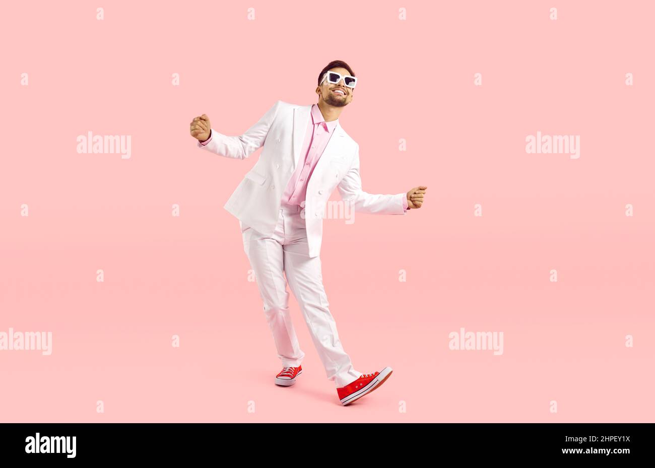 Joyeux drôle jeune homme en costume blanc, baskets et lunettes dansant sur fond rose Banque D'Images