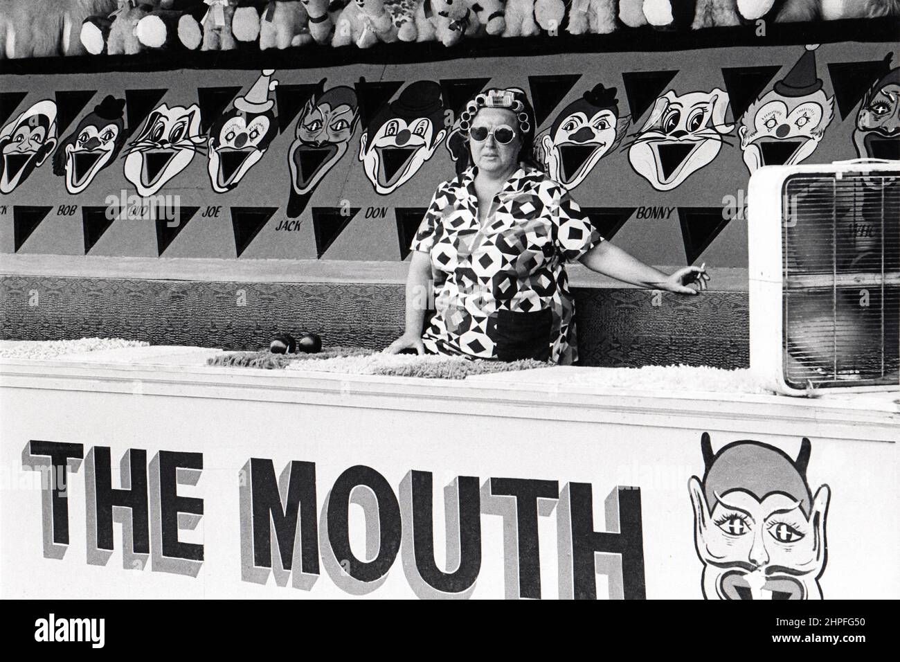 Un opérateur de jeux d'arcade fume une cigarette, porte des cache-boucles et se tient derrière un panneau qui dit « la bouche ». À Coney Island, Brooklyn, New York, vers 1976. Banque D'Images