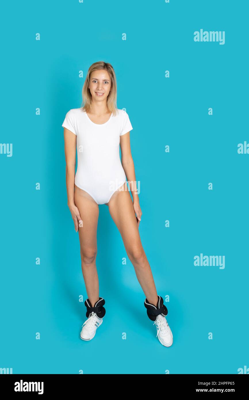 Femme souriante portant un body blanc et des poids de poignet posant regardant la caméra faisant quelques exercices pour être en bonne forme sur fond turquoise Banque D'Images