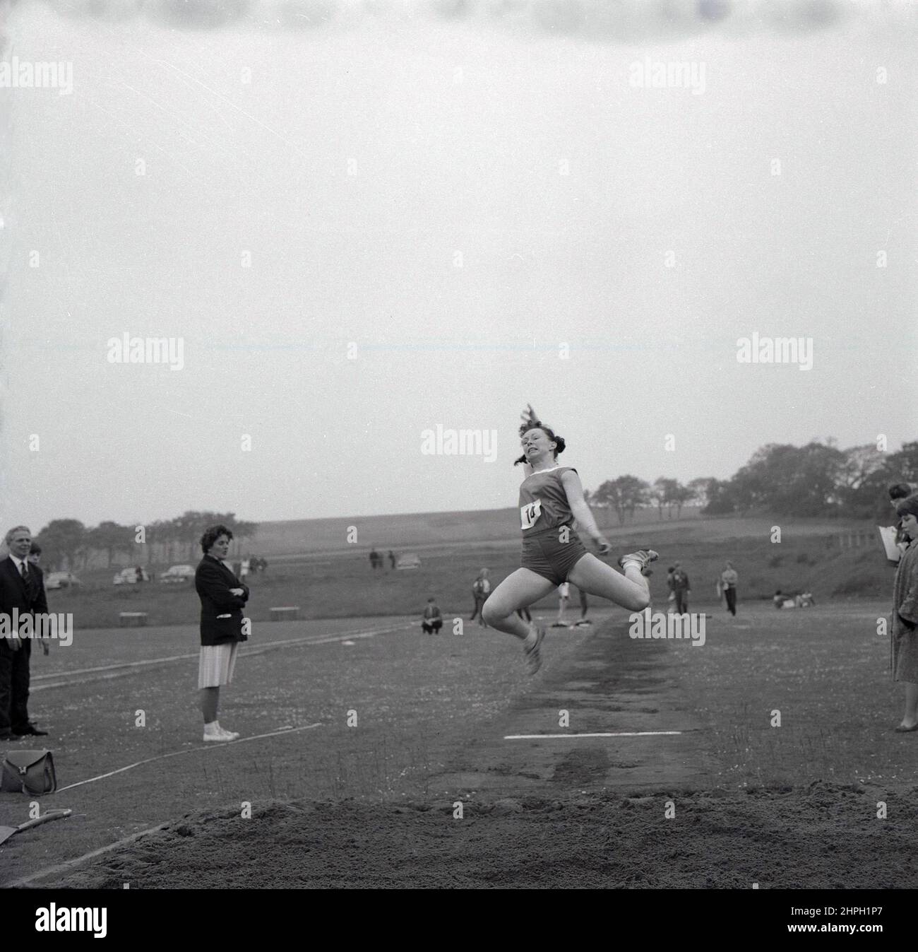 1960s, historique, une athlète féminine à mi-parcours faisant le long saut, Fife, Écosse, Royaume-Uni. La vitesse, la force et l'agilité sont toutes nécessaires dans cet événement sur piste et sur le terrain, où l'objectif est de sauter le plus loin possible d'un point de décollage, où nous voyons ici une dame officielle debout en face. Un événement masculin dans les premiers Jeux Olympiques modernes de 1896, les femmes ont pris part pour la première fois au long saut olympique en 1948 à Londres. Banque D'Images