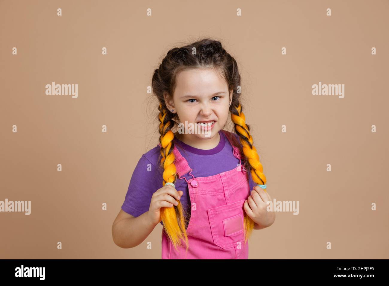 Portrait de la petite colère, femelle tenant des tresses jaunes kanekalon avec les yeux vicieuses regardant l'appareil photo dans la combinaison rose et le t-shirt violet sur beige Banque D'Images