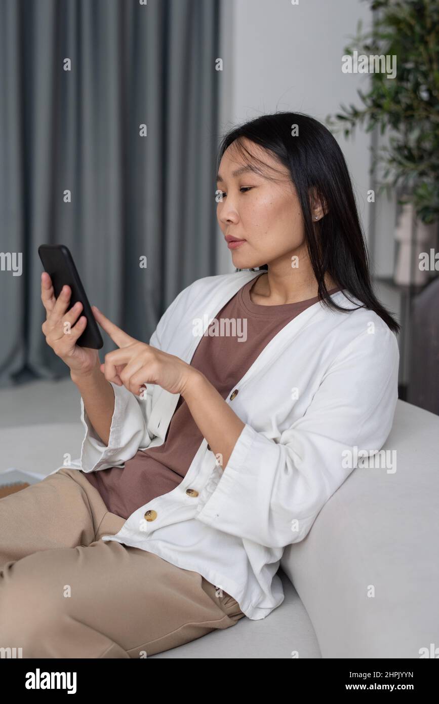 Jeune femme asiatique contemporaine indépendante ou employée défilant dans un smartphone tout en étant assise sur un canapé à la maison et travaillant à distance Banque D'Images