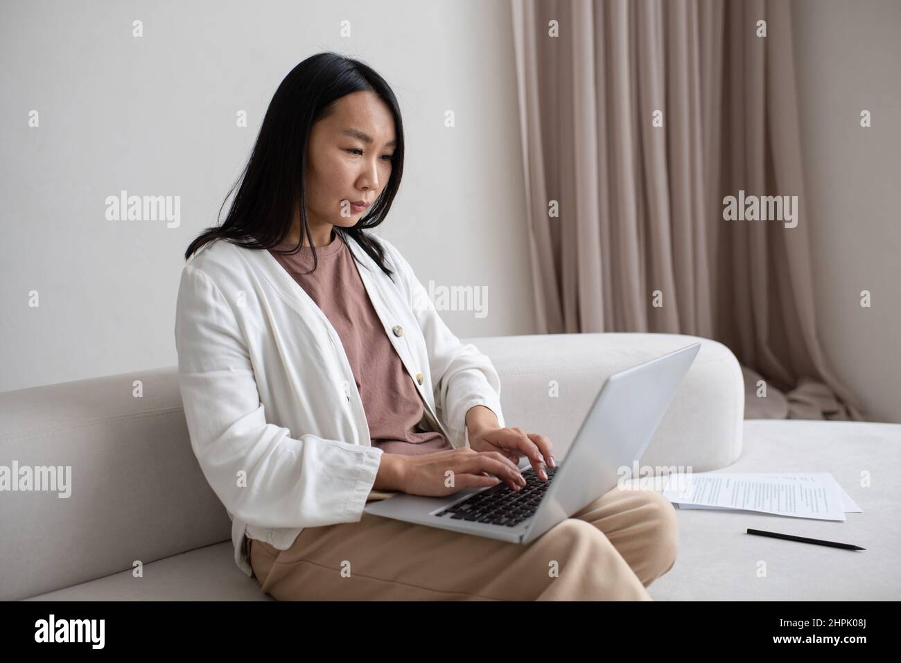 Jeune femme asiatique sérieuse avec un ordinateur portable à genoux, se concentrant sur le réseau tout en étant assise sur un canapé et en travaillant de chez elle Banque D'Images