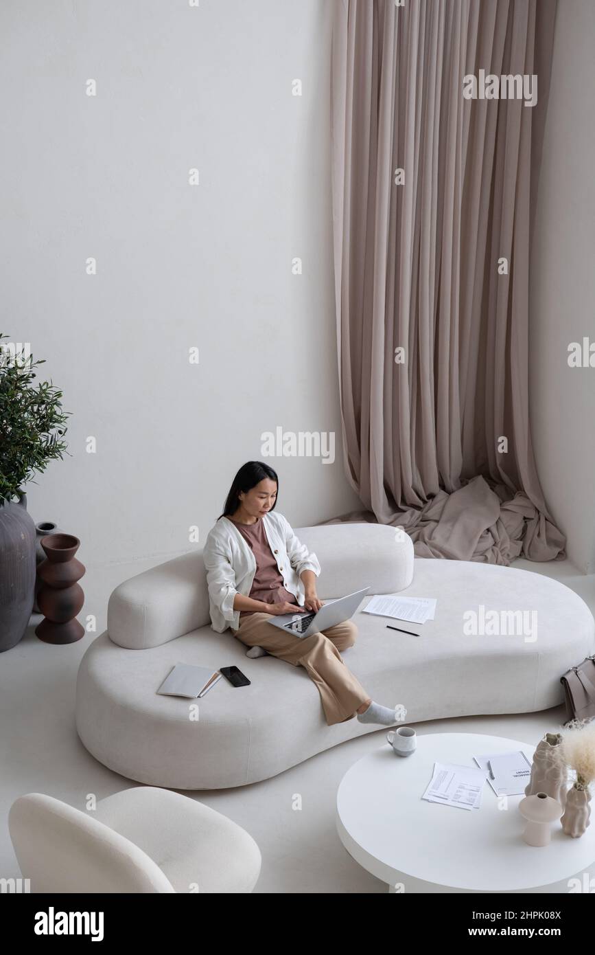 Jeune femme employée ou indépendante avec ordinateur portable assis sur un canapé blanc confortable tout en réseau dans le grand salon de l'appartement moderne Banque D'Images