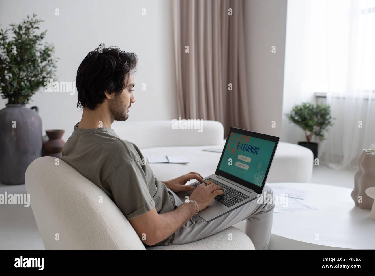Jeune homme contemporain avec un ordinateur portable à genoux prenant le cours en ligne de l'étude tout en s'asseyant sur blanc confortable canapé dans le grand appartement Banque D'Images