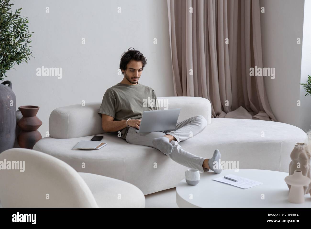 Jeune homme en vêtements décontractés assis sur un canapé et dactylographiant sur un clavier d'ordinateur portable tout en analysant les données financières en ligne Banque D'Images