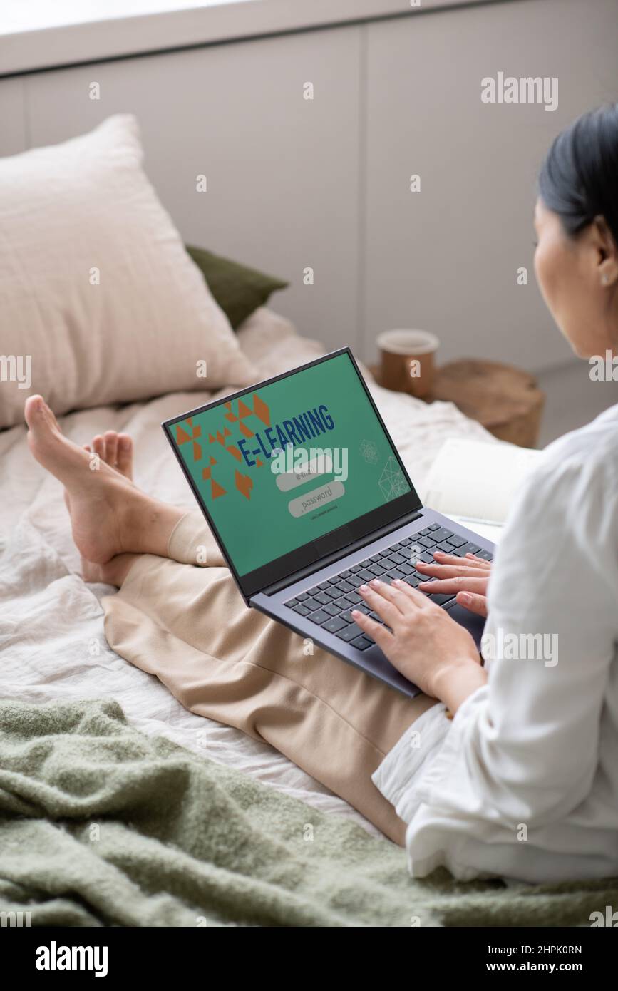 Jeune femme contemporaine en vêtements décontractés assise sur le lit et dactylographiant sur le clavier d'ordinateur portable pendant la leçon en ligne tout en étudiant à distance Banque D'Images
