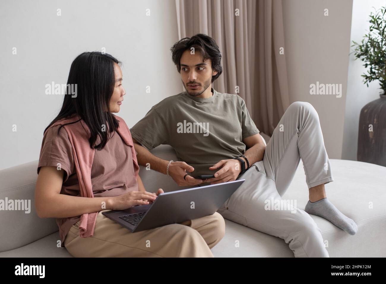 Jeune homme d'affaires confiant avec un ordinateur portable parlant à une femme asiatique tout en étant assis sur un canapé et en discutant de données en ligne Banque D'Images
