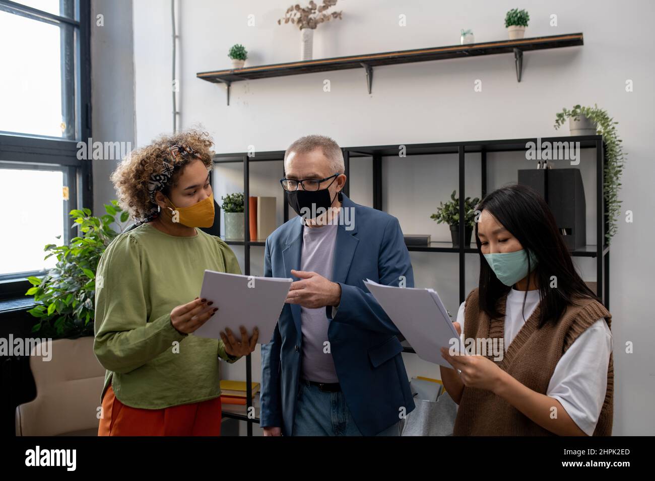 Homme d'affaires mature dans le masque pointant sur le document pendant la discussion avec une jeune collègue biracial lors de la réunion de travail au bureau Banque D'Images