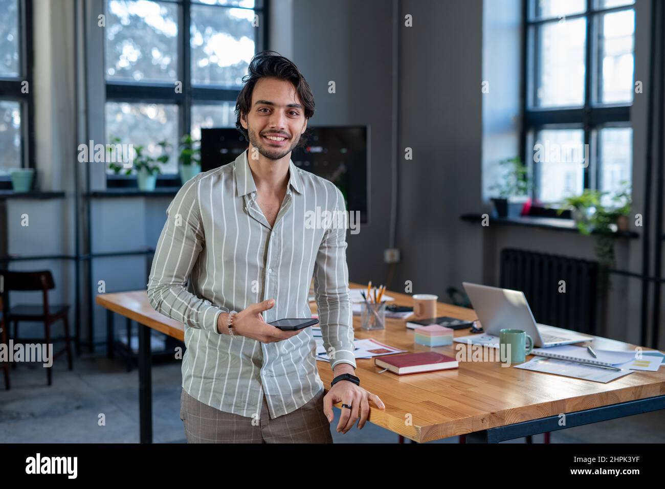 Jeune employé souriant dans une chemise rayée décontractée debout près du lieu de travail contre table avec des papiers financiers et d'autres fournitures Banque D'Images