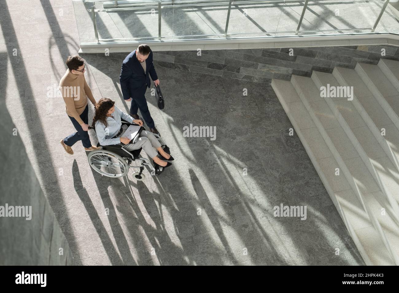 Vue ci-dessus de deux hommes d'affaires marchant par une jeune femme en fauteuil roulant tout en l'aidant à se déplacer à l'intérieur du centre de bureau Banque D'Images