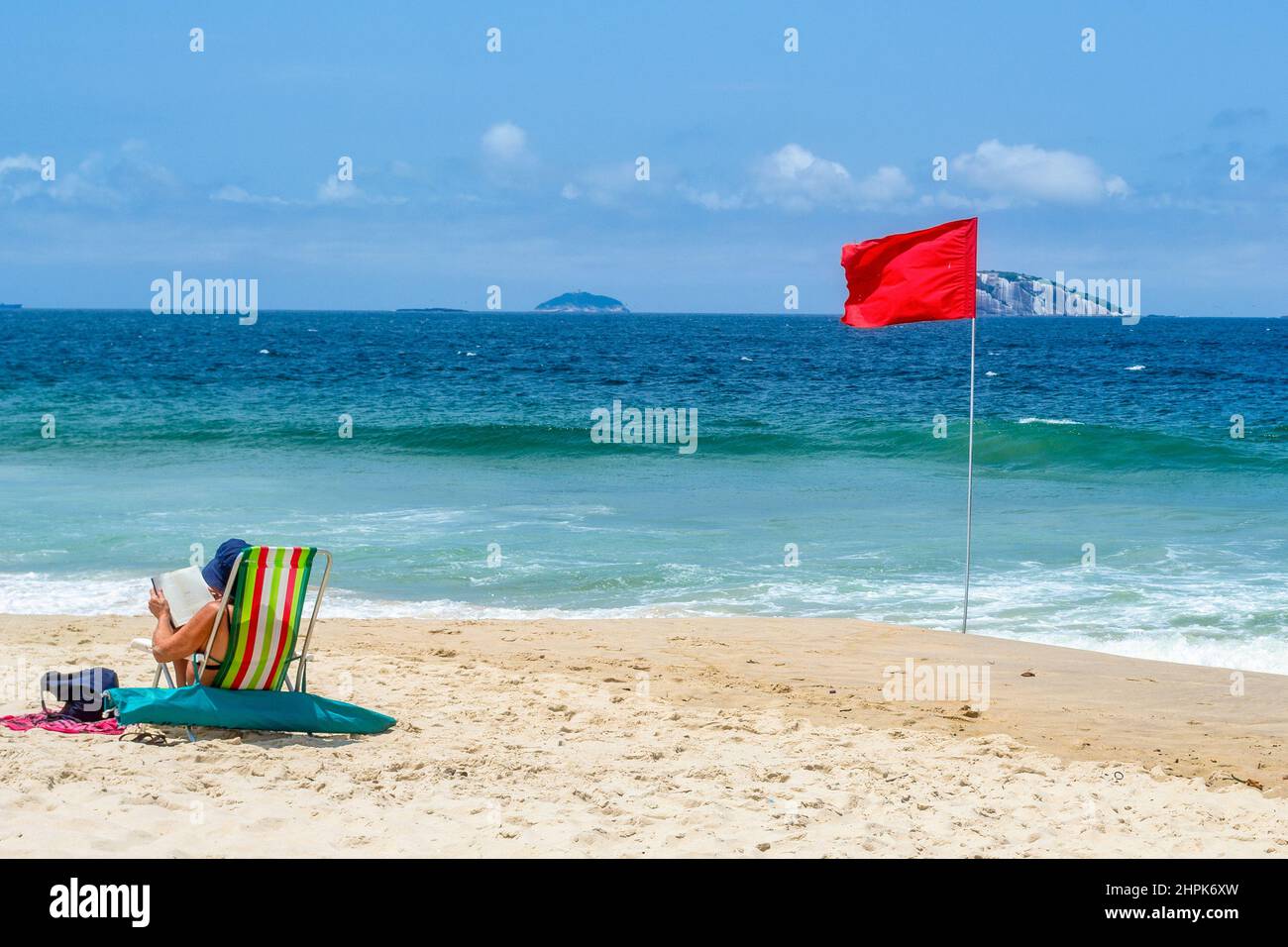 Un touriste dans une chaise dans le sable par un drapeau rouge. Ipanema Beach est un endroit célèbre et une destination de voyage majeure dans le pays sud-américain. Banque D'Images