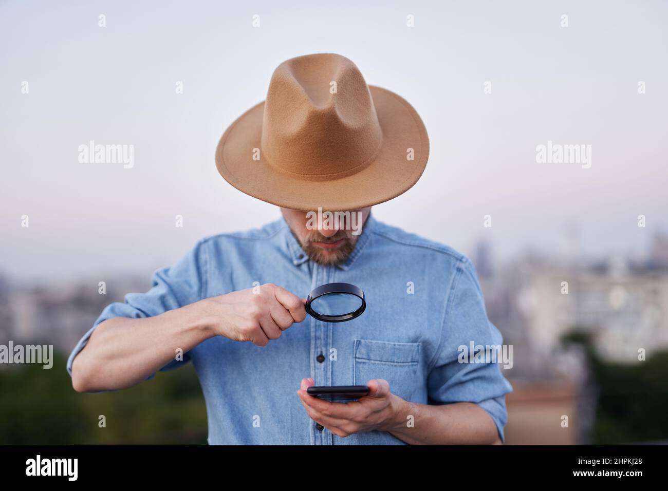 Homme barbu regardant avec la loupe sur le téléphone portable. Homme portant un Jean-shirt et un chapeau regardant avec la loupe au smartphone comme détective, enquêteur ou concept de recherche. Image de haute qualité Banque D'Images