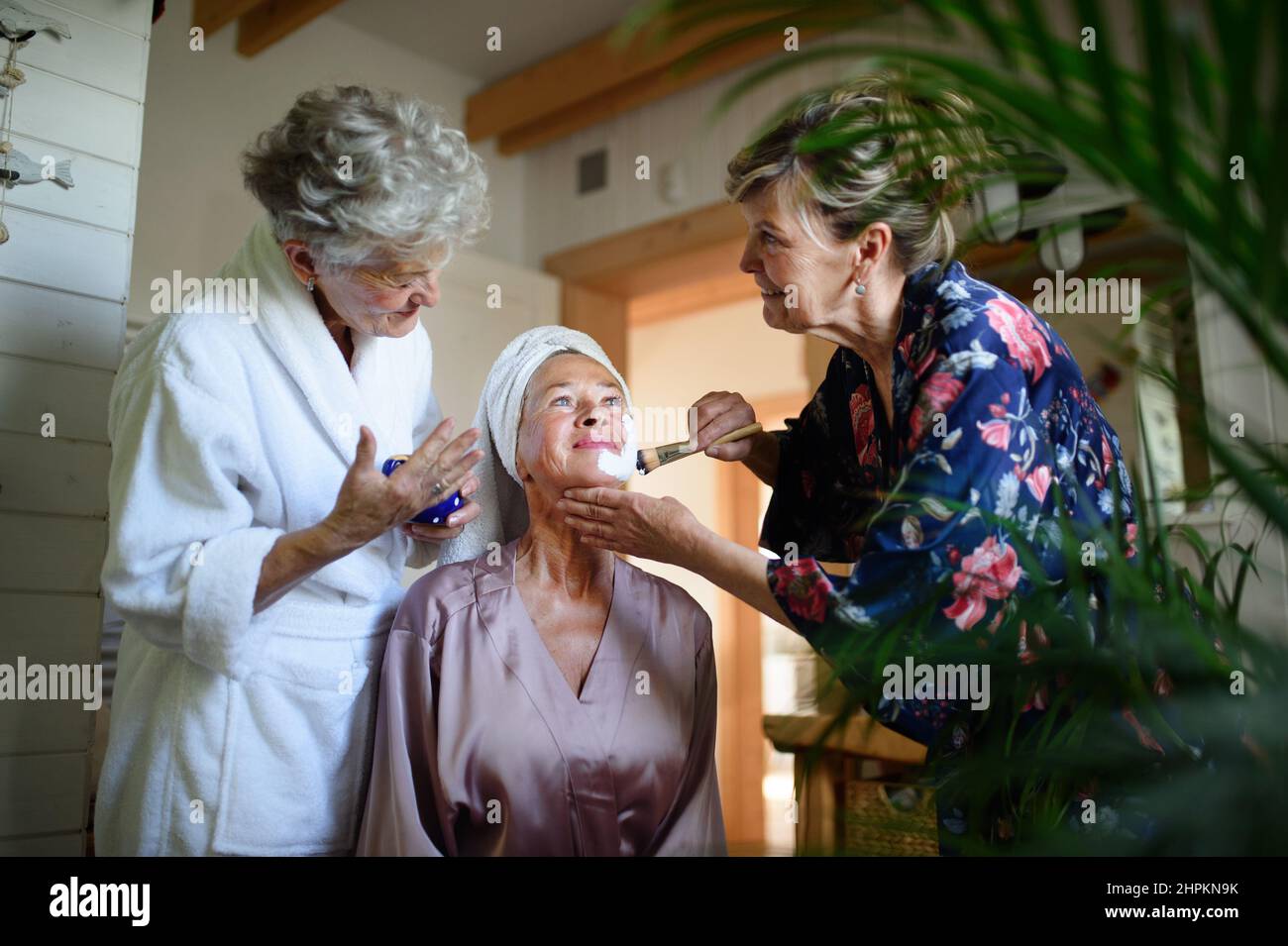 Des femmes âgées heureuses dans des peignoirs appliquant un masque à l'intérieur dans la salle de bains, concept d'autosoin. Banque D'Images