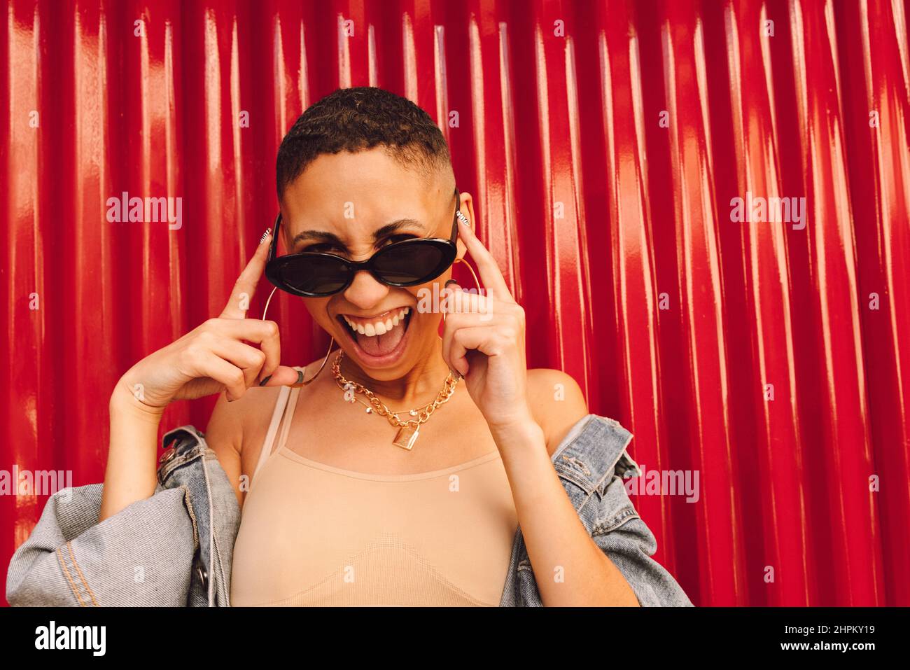 Jeune femme gaie souriant à l'appareil photo tout en touchant ses lunettes de soleil. Bonne jeune femme debout sur fond rouge. Jeune femme se sentant vib Banque D'Images