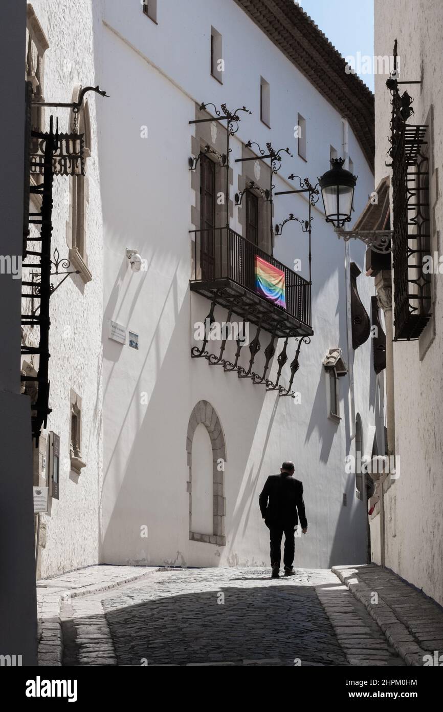 L'homme marche dans une rue étroite de Sitges, en Espagne. Vue arrière. Lumière du soleil frappant la façade du bâtiment. Drapeau de la communauté LGBT suspendu sur une terrasse. Banque D'Images