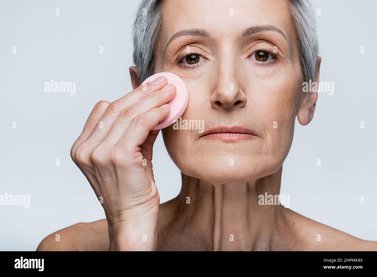femme mature tenant une éponge de beauté et appliquant le fond de teint de maquillage isolé sur gris Banque D'Images