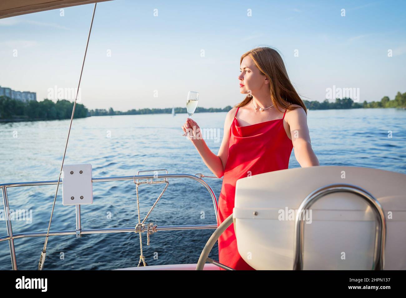 L'élégante femme dans une longue robe rouge tenant un verre de champagne debout sur un yacht et à la recherche d'une vue sur le coucher du soleil sur la rivière Banque D'Images