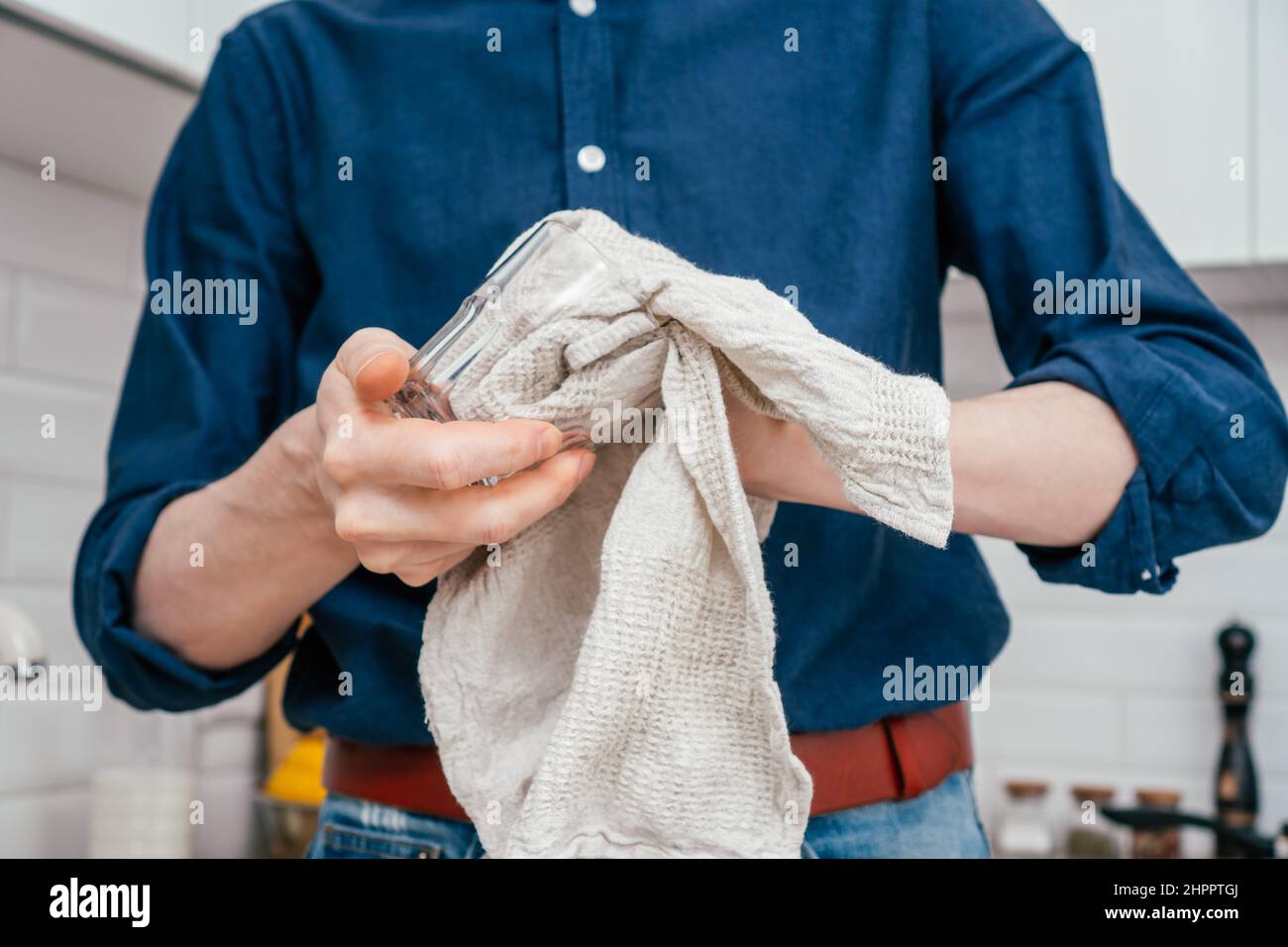 Gros plan de l'homme méconnaissable en chemise bleue, jeans et ceinture en cuir rouge verre séchant avec coton gris serviette de cuisine debout dans blanc brillant moderne Banque D'Images