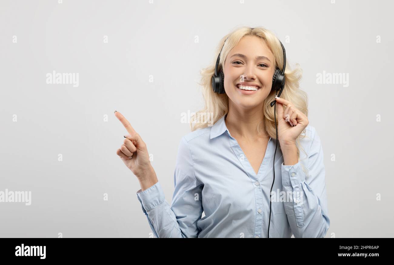 Jeune femme heureuse opérateur de centre d'appels portant un casque et pointant de côté sur l'espace libre, le fond clair, le panorama Banque D'Images