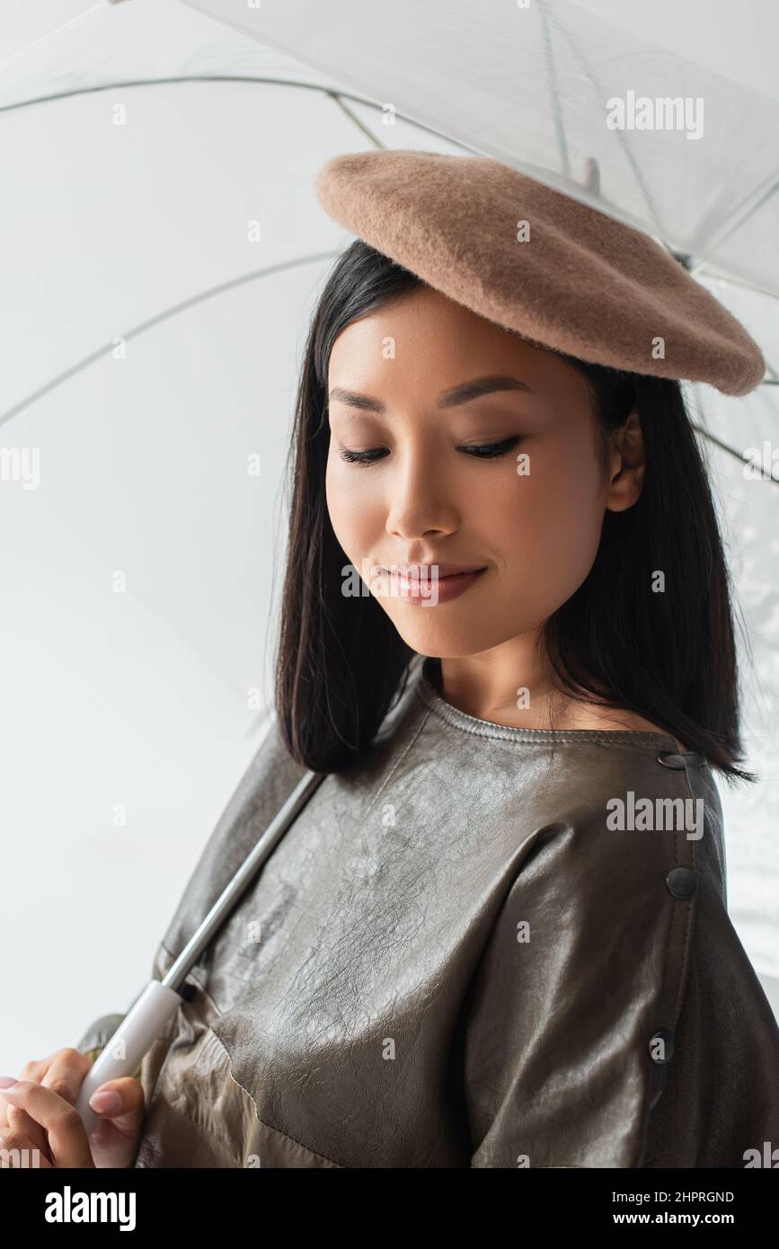 jolie femme asiatique en béret beige souriant sous un parapluie transparent isolé sur gris Banque D'Images