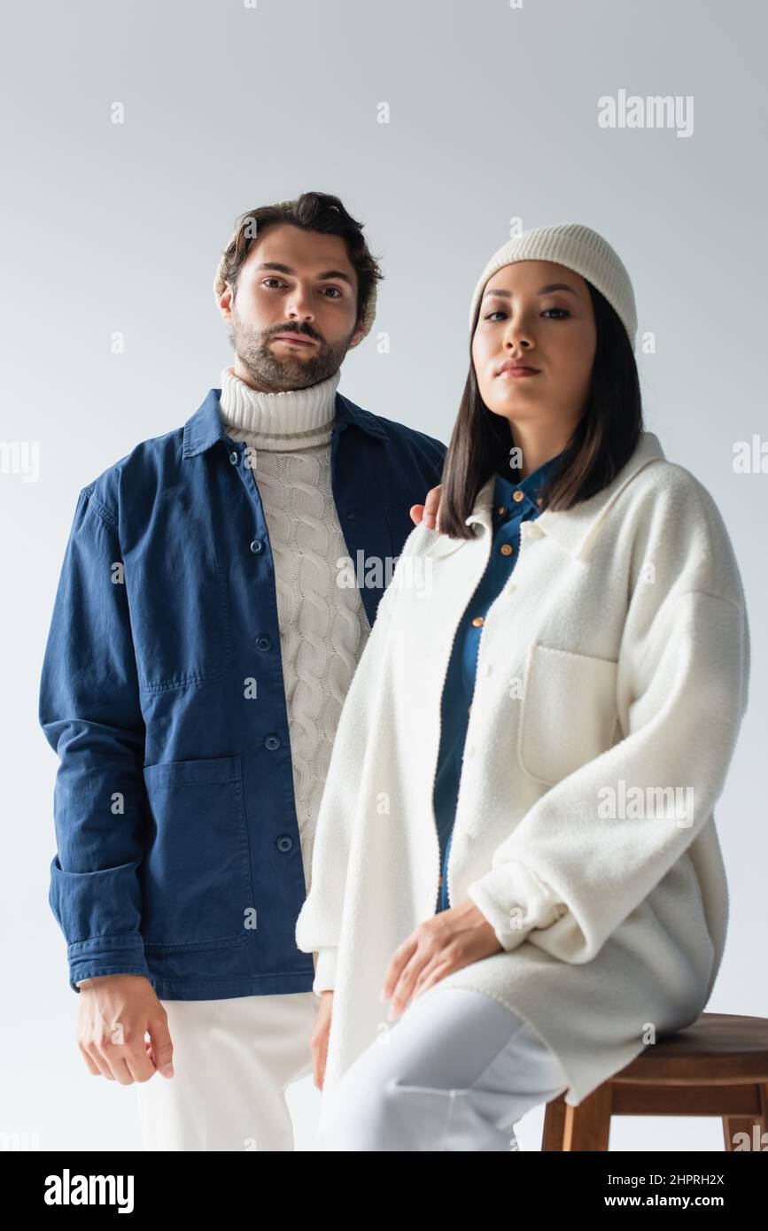 homme tendance regardant l'appareil photo près d'une femme asiatique en blouson blanc et bonnet isolé sur gris Banque D'Images