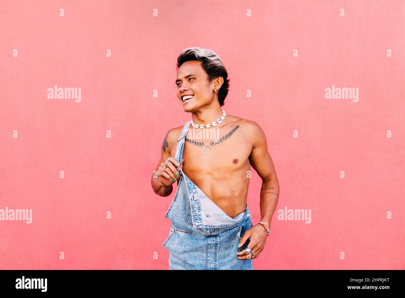 Portrait d'un jeune homme souriant dans une combinaison en denim qui regarde loin tout en se tenant sur un mur rose Banque D'Images
