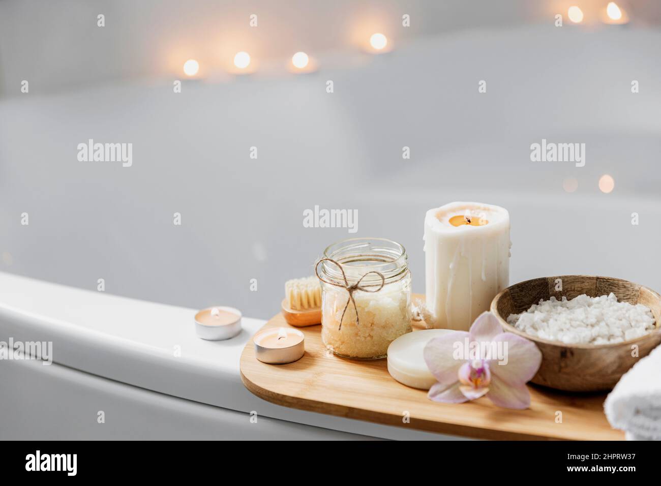 Préparation pour le traitement de spa de l'hôtel ou la procédure de bain à la maison. Lavabo blanc dans la salle de bains avec accessoires sur le plateau. Bougies allumées, débarbouillette, savon, brosse à pied, bouteille avec sel de mer, fleur d'orchidée Banque D'Images