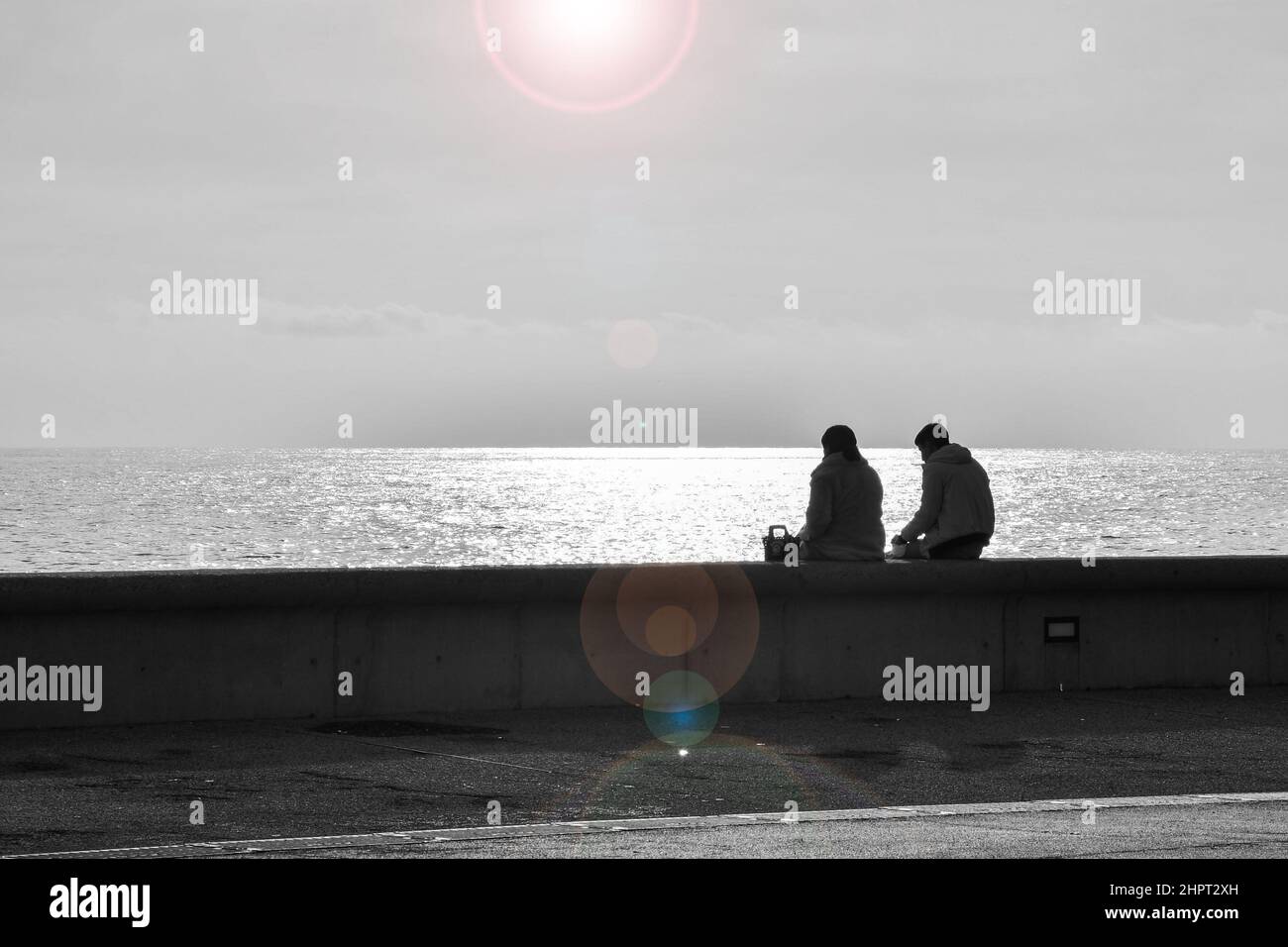 Vue à contre-jour d'un couple assis sur un mur bas sur le front de mer de Sanremo avec des fusées éclairantes, Imperia, Ligurie, Italie Banque D'Images