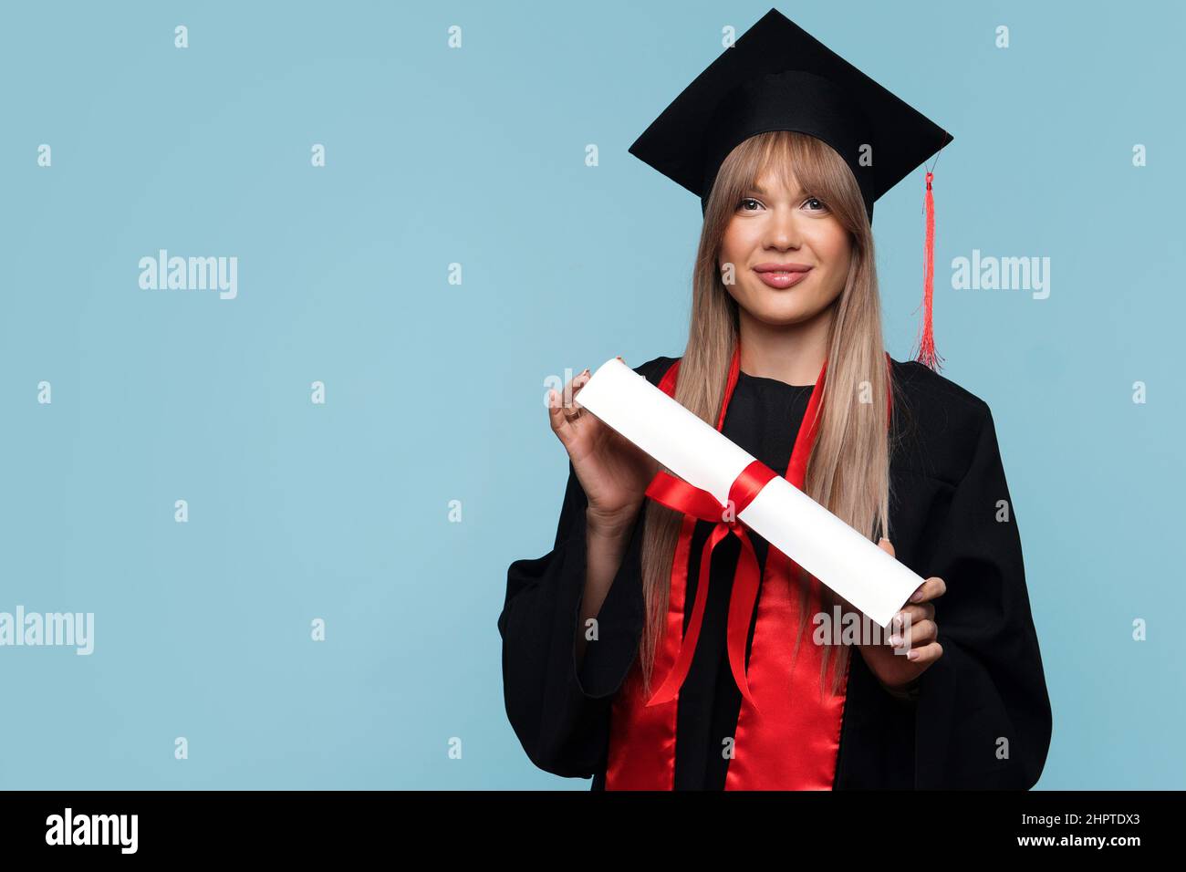 Jeune fille diplômé chapeau avec diplôme sur fond bleu clair. Jeune femme  blonde portant une casquette de remise des diplômes et une robe de  cérémonie tenant le certificat attaché avec un ruban