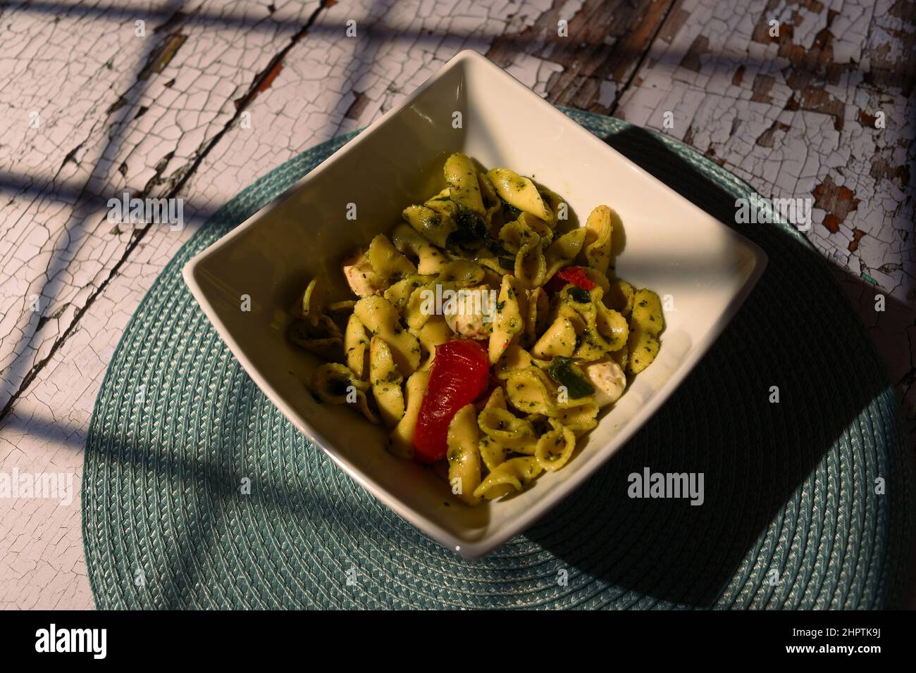 Salade de pâtes à la mozzarella, aux tomates et au pesto dans un plat carré blanc sur une table en difficulté avec des ombres de fenêtre tombant sur la table et le plat. Banque D'Images