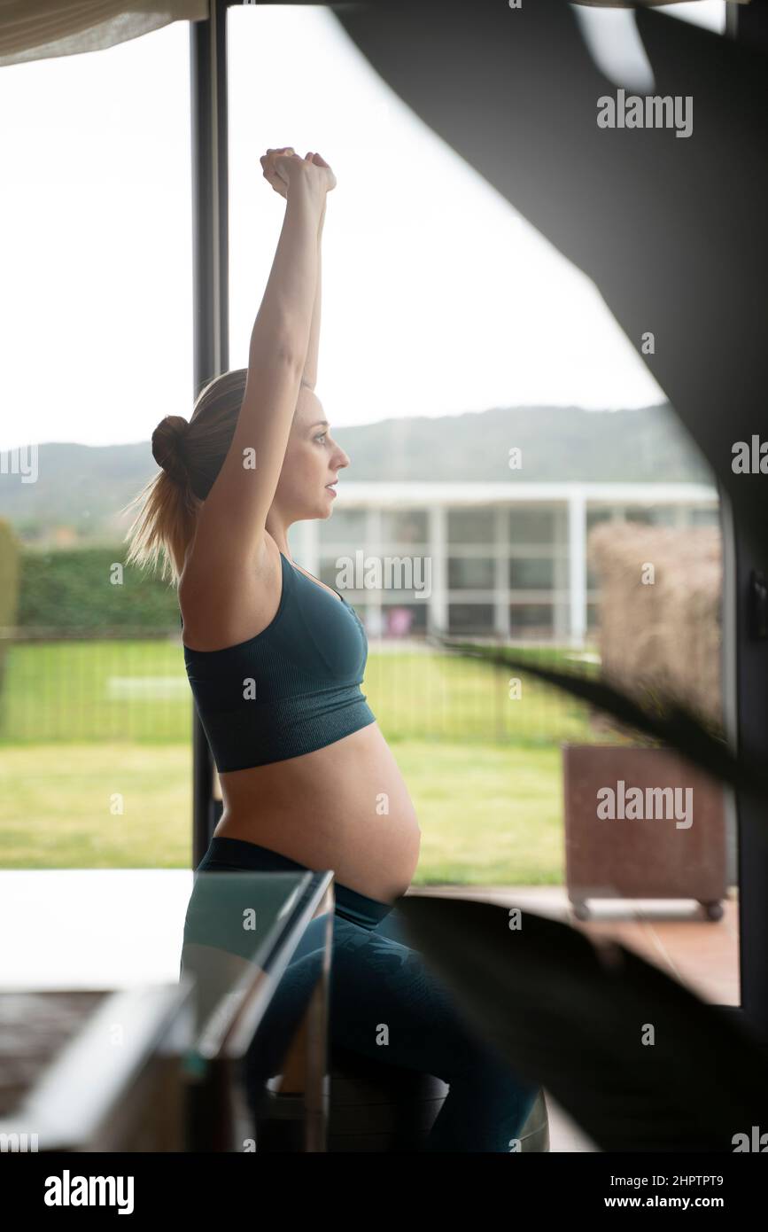 Femme enceinte blonde pratiquant le yoga dans le salon à côté des fenêtres Banque D'Images