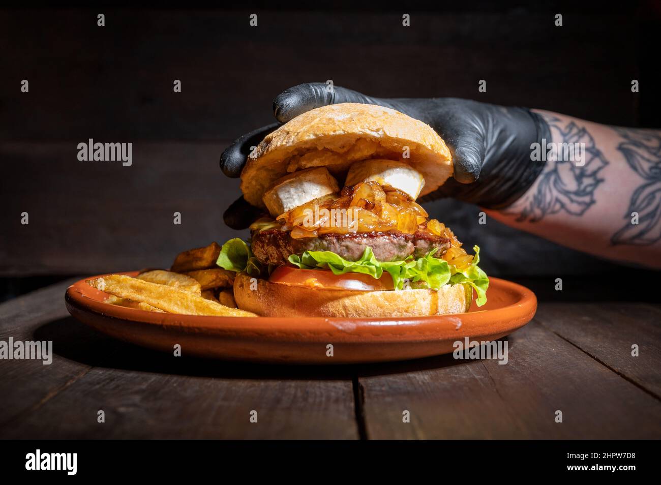 Gros plan d'un chef méconnu qui termine un hamburger de bœuf de qualité supérieure avec du fromage de chèvre, des oignons caramélisés, servi avec des frites sur une assiette rustique sur une table en bois. Photographie de haute qualité Banque D'Images