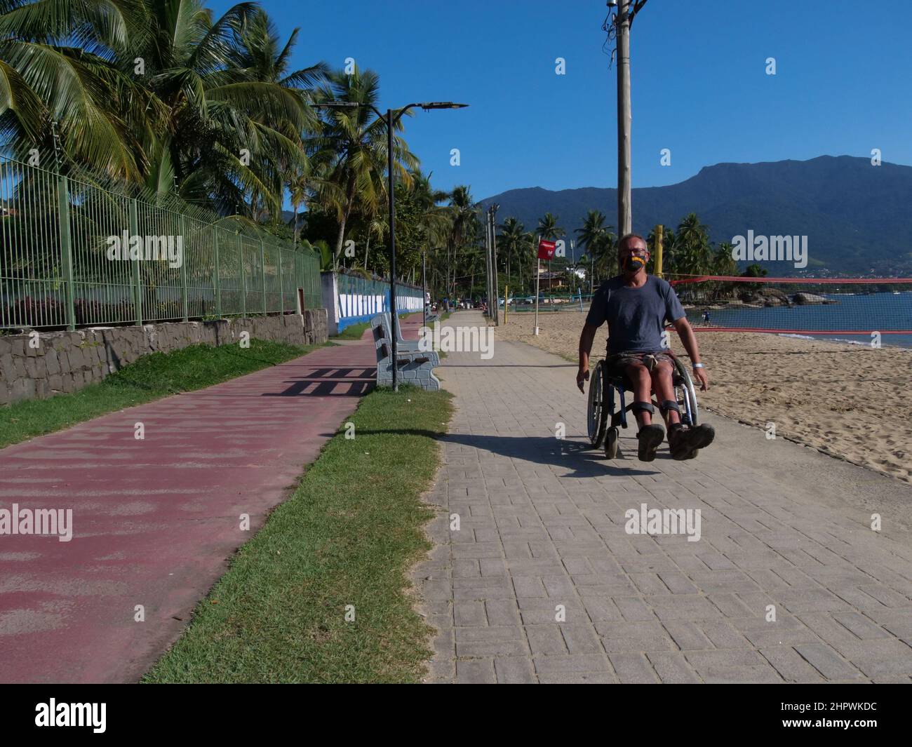 Un homme handicapé en fauteuil roulant s'approche du spectateur. Il porte un masque facial conformément aux réglementations Covid-19 existantes. Banque D'Images