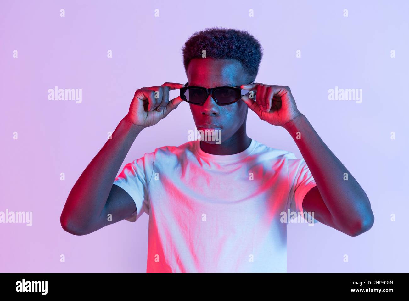 Modèle africain américain sérieux en tant que t-shirt debout et réglage de lunettes de soleil noires en studio avec éclairage coloré Banque D'Images