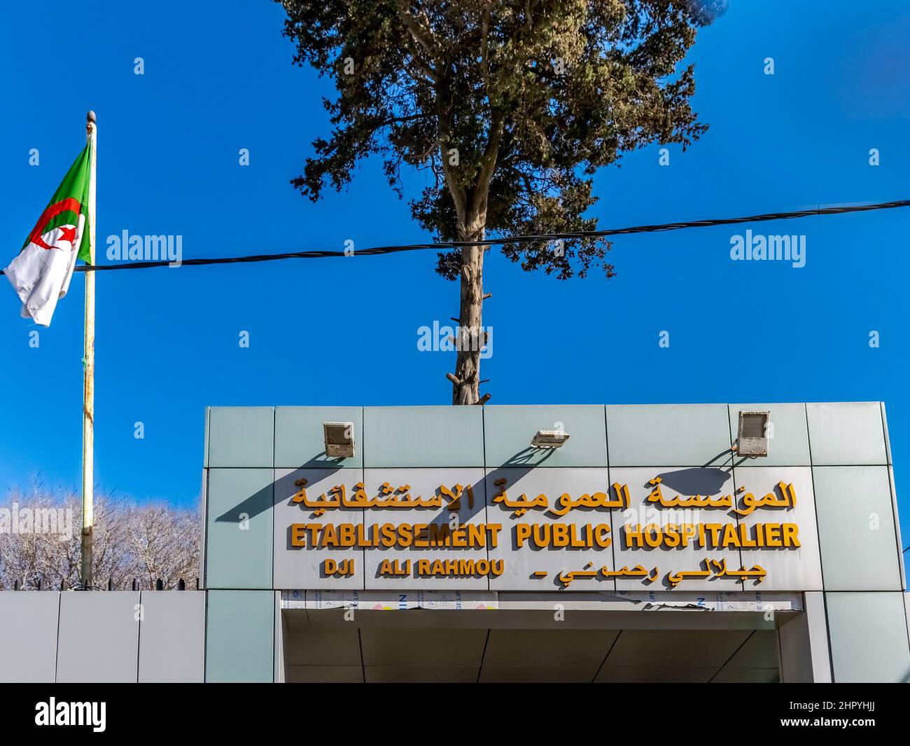 Centre hospitalier public Rahmouni Djillali entrée. Nommez la plaque en arabe et en français. Drapeau algérien, câble électrique, arbres, ciel bleu. Banque D'Images