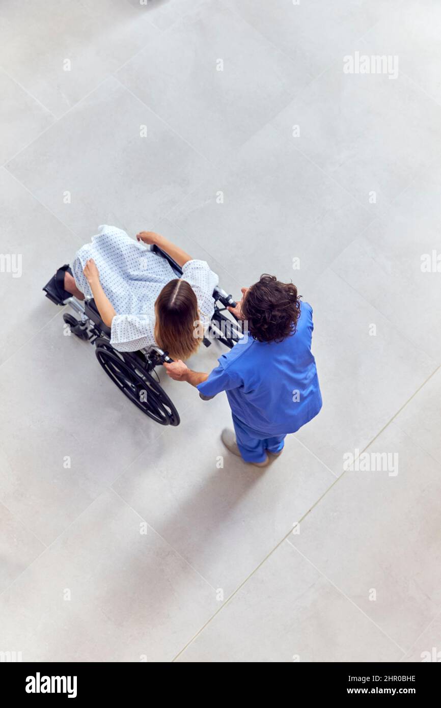 Un coup de tête d'une infirmière portant des produits de nettoyage poussant une patiente en fauteuil roulant dans le bâtiment de l'hôpital Banque D'Images