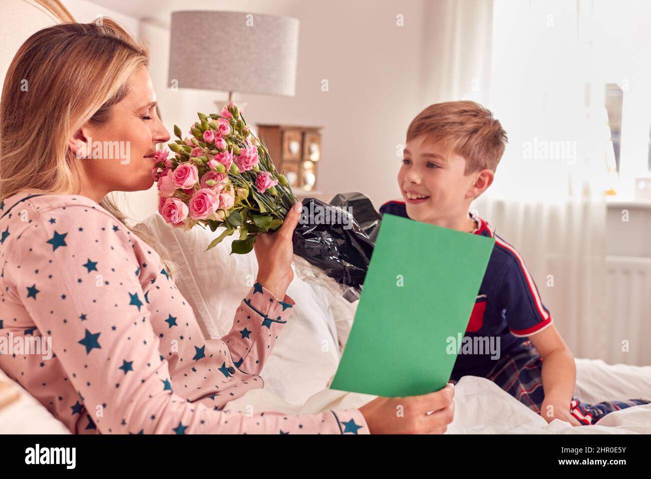 Fils aimant mère surprenante au lit avec fleurs et carte maison à l'anniversaire ou à la fête des mères Banque D'Images