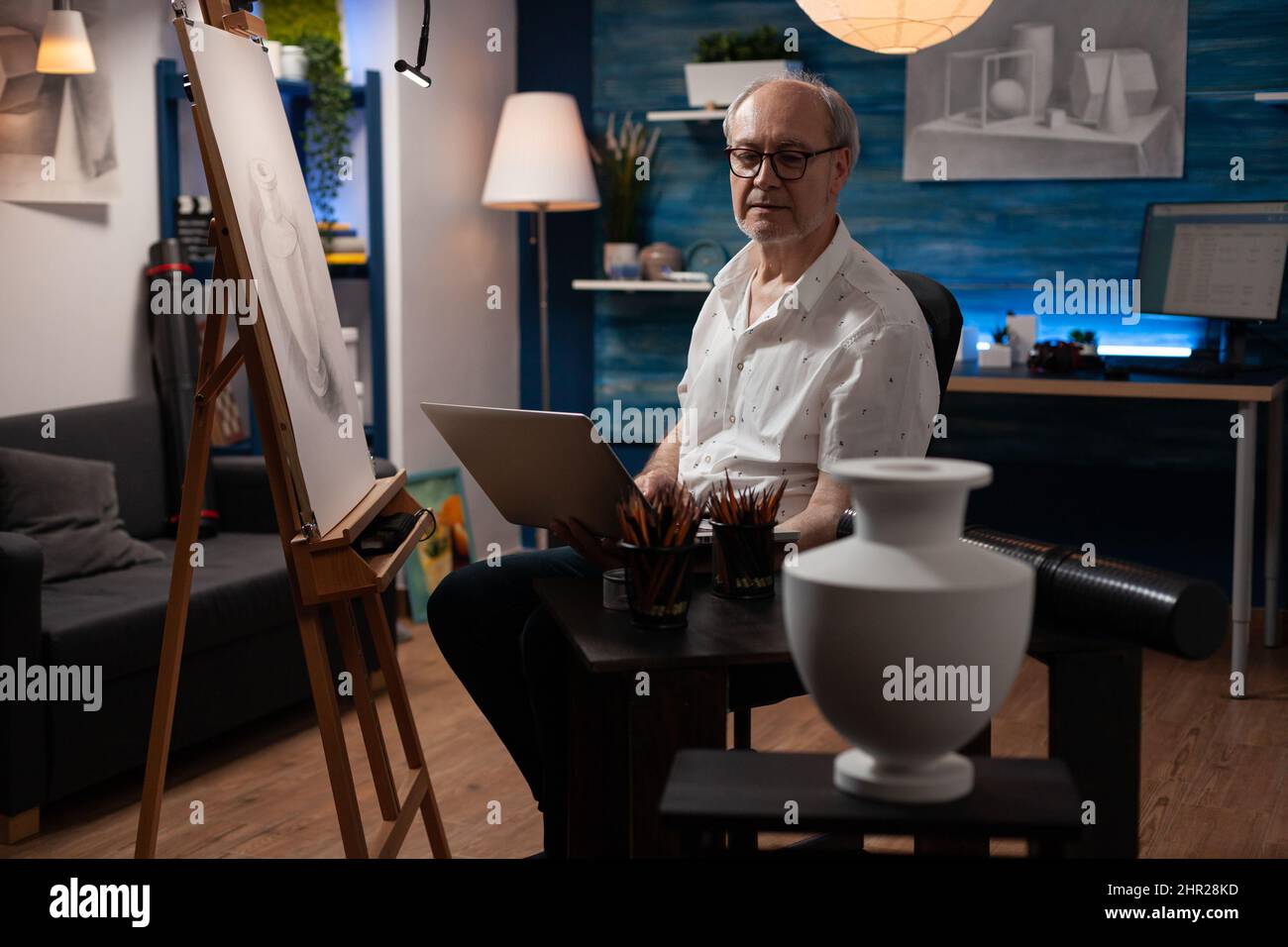 Artiste senior tenant un ordinateur portable assis dans un studio d'art avec des outils de dessin professionnels regardant le vase modèle. Homme à la retraite qui trouve son inspiration en ligne sur les réseaux sociaux devant un chevalet avec croquis. Banque D'Images