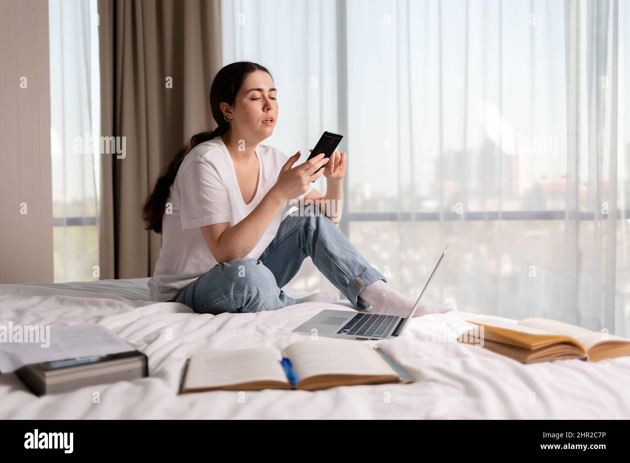 Une jeune femme de race blanche est assise sur un lit et utilise un smartphone. Copier l'espace. Le concept de l'éducation en ligne et la dépendance aux réseaux sociaux. Banque D'Images