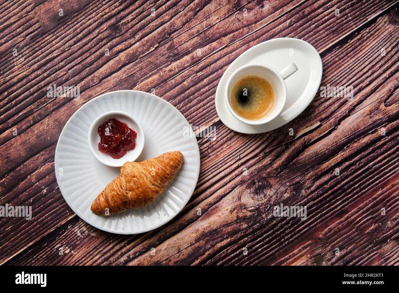 Un café croissant, de la confiture de fraises et de l'espresso sur une assiette ronde texturée blanche sur un fond de bois pris d'en haut, un favori de petit déjeuner dans le monde entier. Banque D'Images