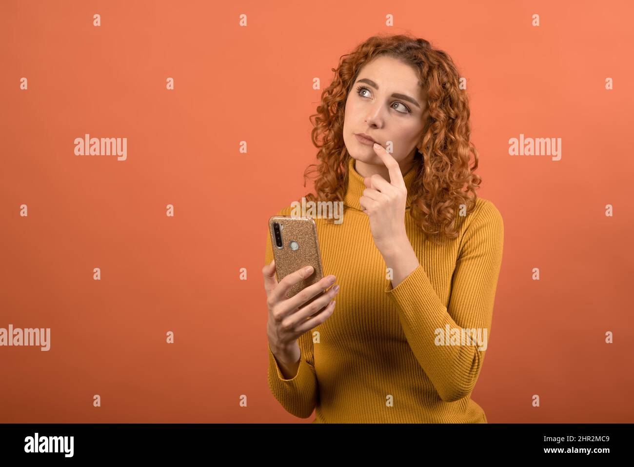 Jeune et attrayante fille caucasienne à tête rouge en orange en utilisant un smartphone avec un aspect pensif isolé sur fond orange studio. Banque D'Images