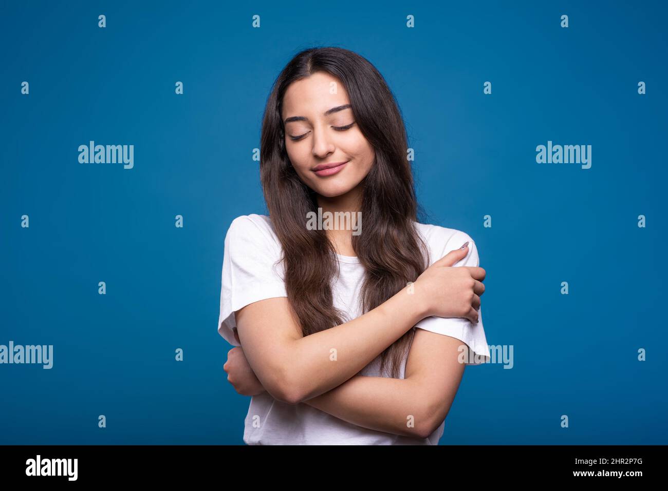 Jolie fille caucasienne ou arabe brune en t-shirt blanc avec les yeux fermés se embrassant isolée sur fond bleu studio. Banque D'Images