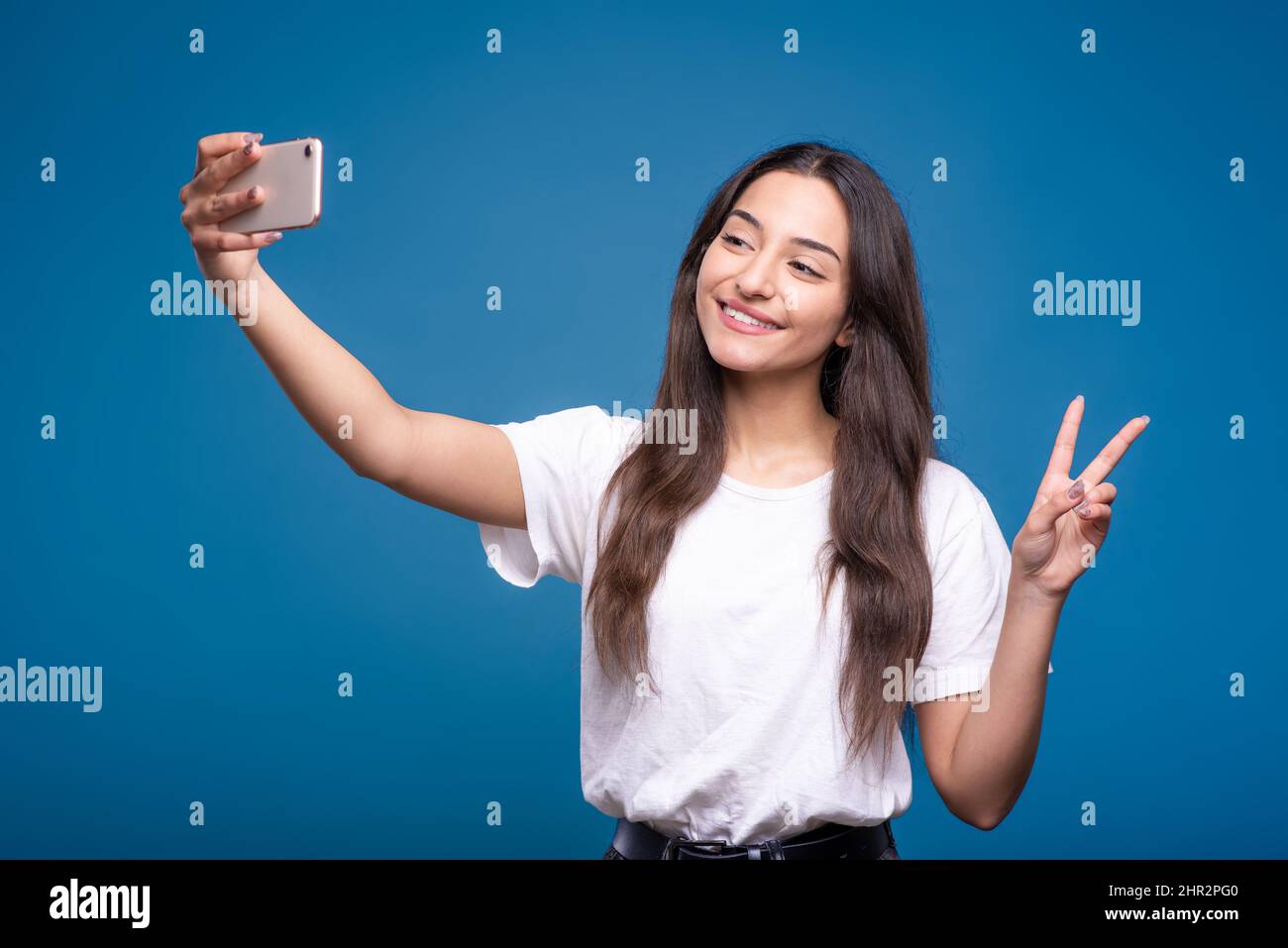 Jolie fille brune caucasienne ou arabe dans un t-shirt blanc prenant un selfie sur un appareil photo de téléphone portable et montrant un geste de paix isolé sur un bleu Banque D'Images