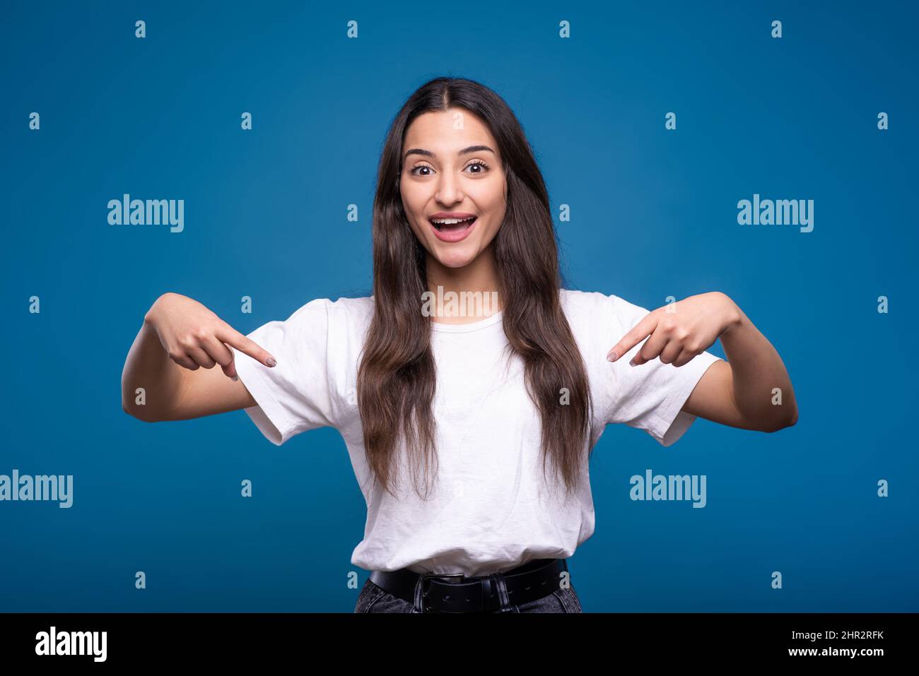 Jeune et attrayante caucasien ou arabe brune fille en t-shirt blanc pointant les doigts vers le bas à l'espace de copie pour la publicité isolée sur le studio bleu dos Banque D'Images