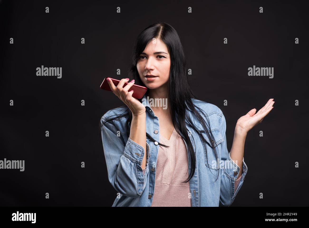 Jolie fille brune caucasienne dans une chemise tenant un smartphone dans sa main et enregistrant un message vocal isolé sur un fond noir de studio. Banque D'Images