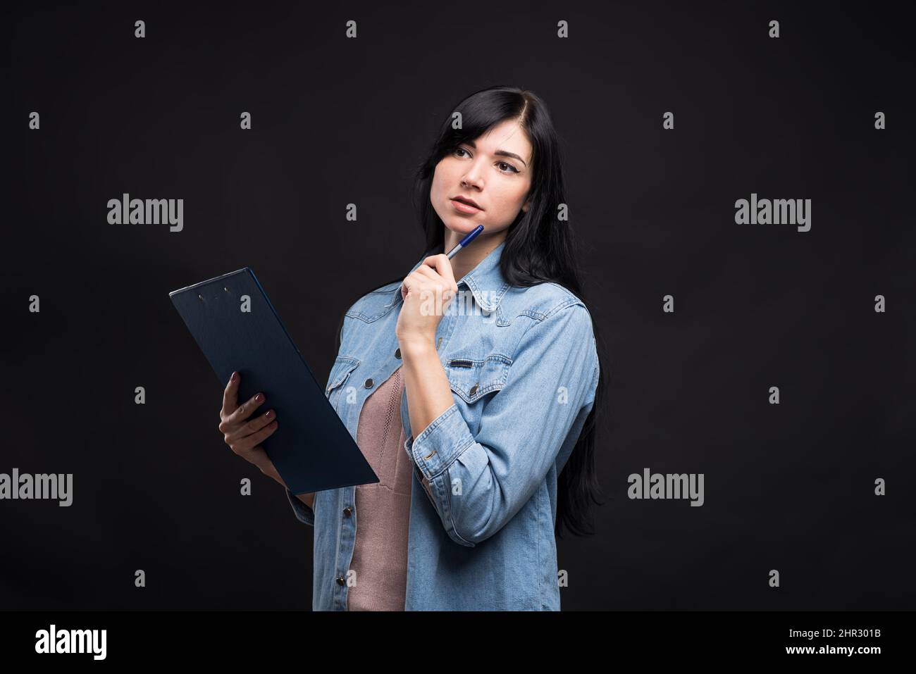 Jolie fille de brune caucasienne dans une chemise tenant une tablette et l'écriture avec un regard pensif isolé sur un fond noir de studio. Banque D'Images