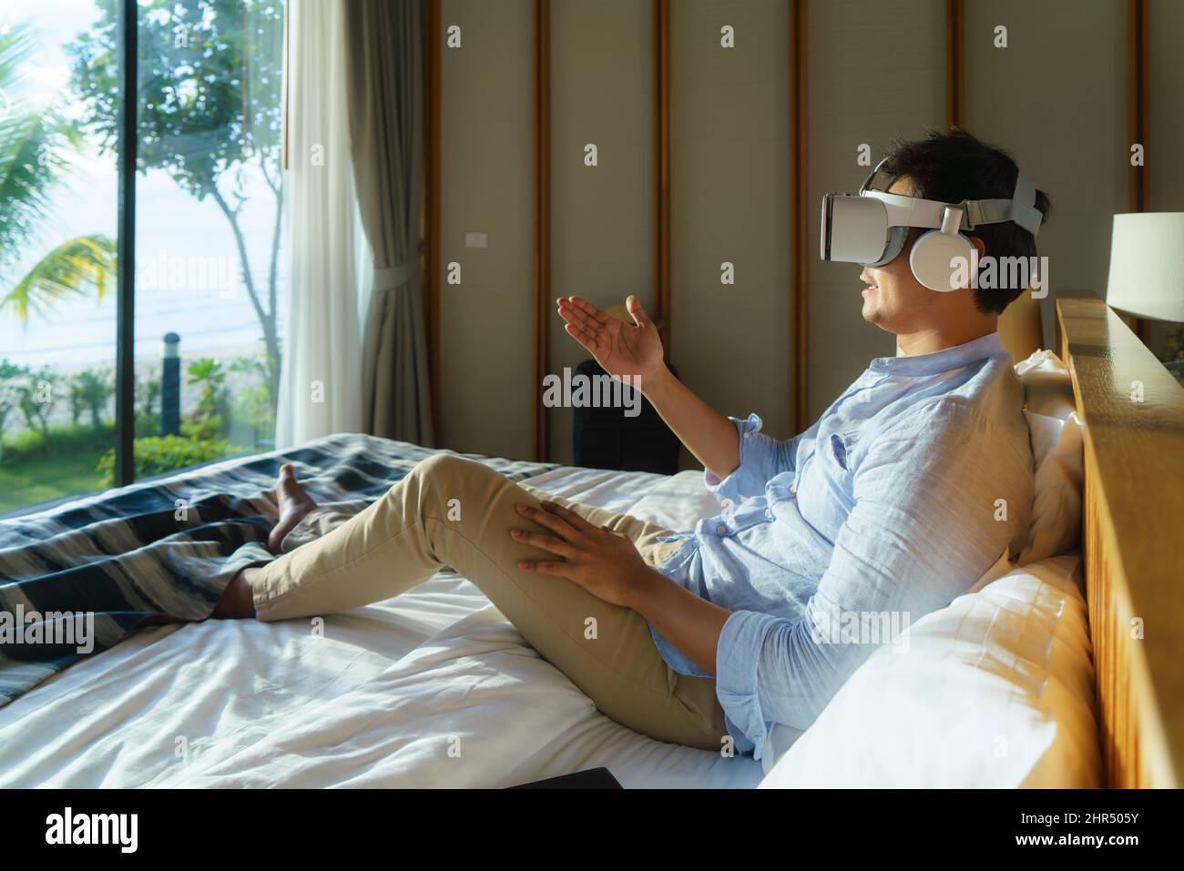 Jeune homme asiatique allongé sur le lit et utilisant des lunettes de réalité virtuelle pour une réunion d'affaires pendant ses vacances d'été de voyage. Banque D'Images