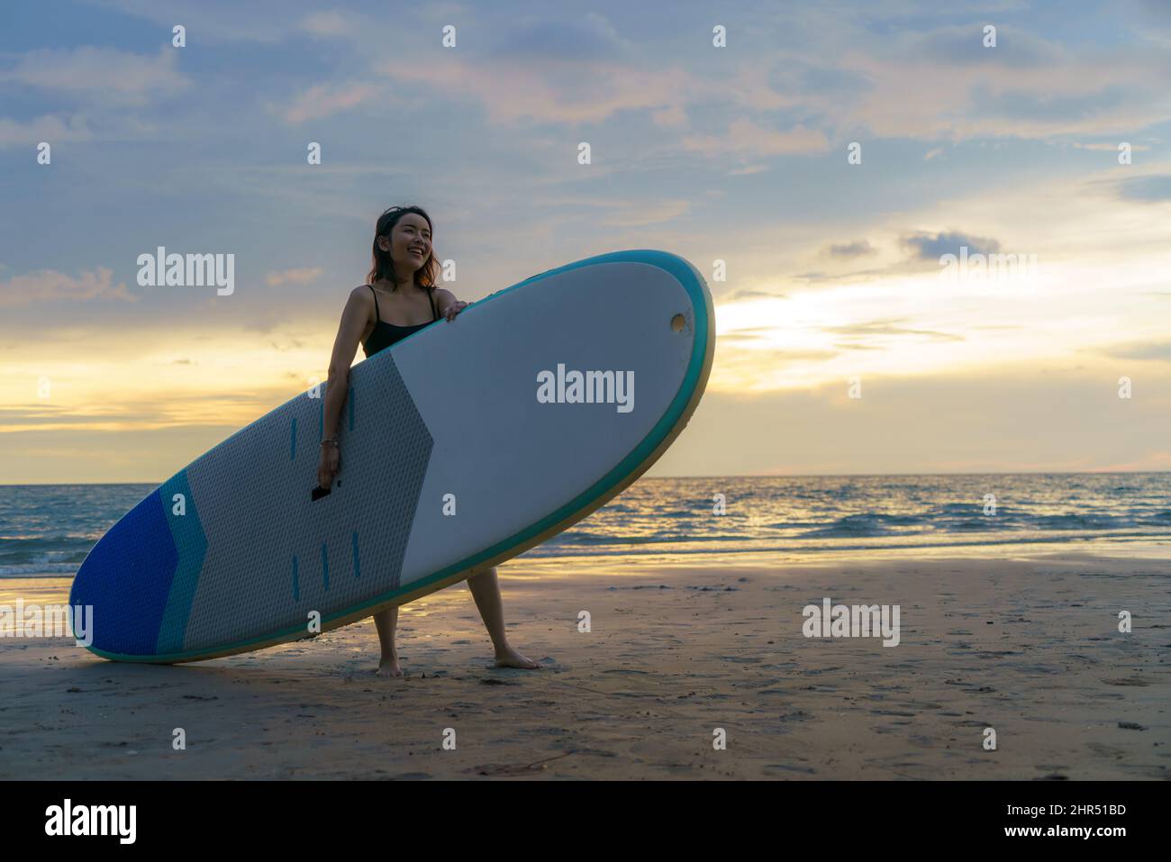 Femme asiatique tenant un panneau supérieur et paddle et marchant sur la plage.En arrière-plan, l'océan et le coucher du soleil.Vue arrière.Surf d'été. Banque D'Images