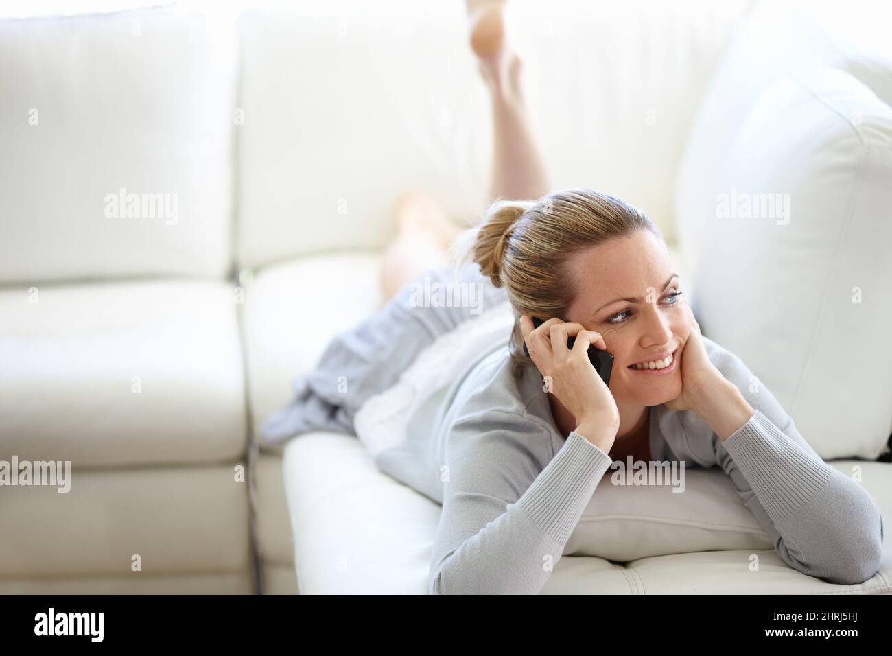 Nous pourrions parler pendant des heures. Une jeune femme heureuse utilisant son téléphone portable tout en étant simplement couché sur un canapé. Banque D'Images